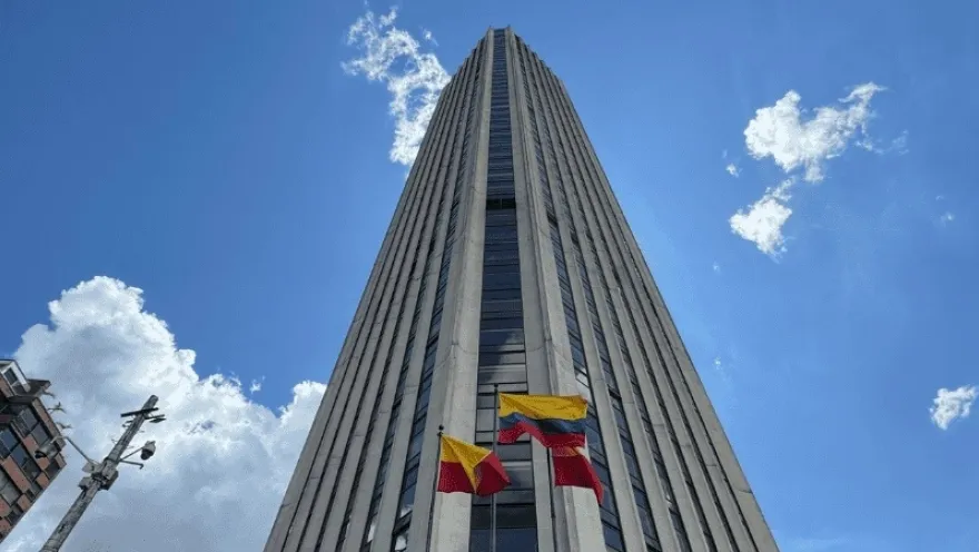 Edificio alto de estilo moderno visto desde abajo, con líneas verticales que acentúan su altura. En la base, varias banderas de Colombia ondean frente a la fachada, bajo un cielo azul con nubes dispersas.