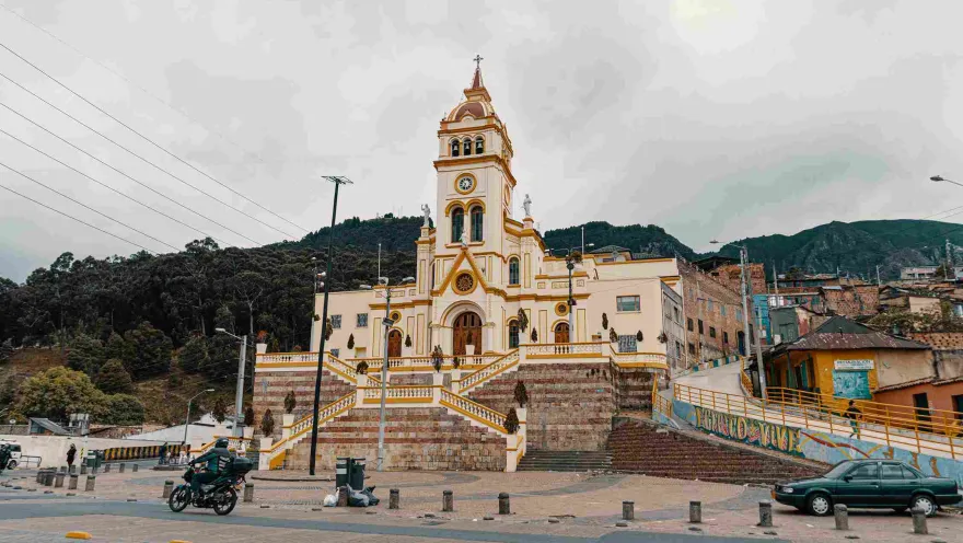 General view of the facade of a light brown church, behind the mountains. In front there is a street with a parked car and a motorcyclist.
