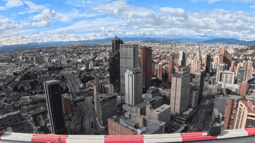 Vista panorámica de una ciudad con numerosos edificios altos y avenidas amplias, observada desde gran altura. El horizonte muestra montañas lejanas bajo un cielo parcialmente nublado.