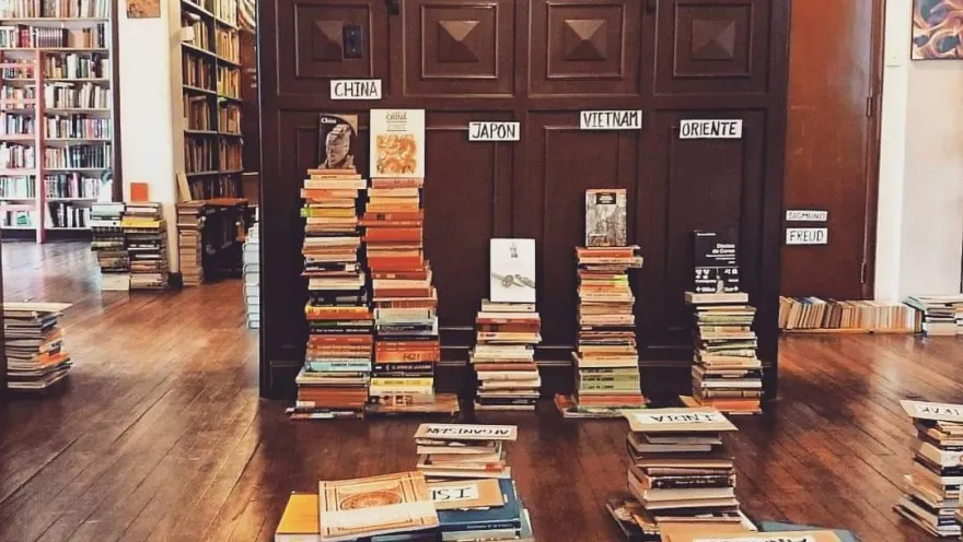 Interior of the bookstore with stacks of books organized by subject on a wooden floor and shelves in the background.