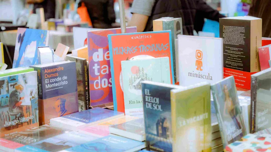 An exhibition table showcasing a diverse selection of books and novels at a literary fair, surrounded by visitors and independent publishers in a vibrant, creative atmosphere.
