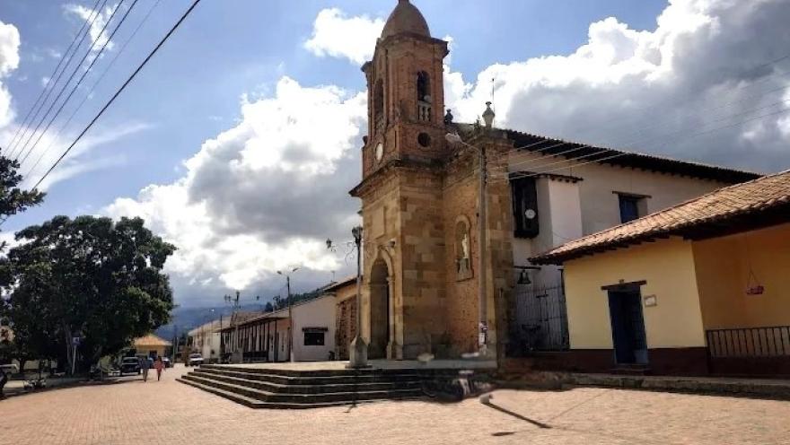 Town church with a stone tower and bell tower, located next to a large plaza, with colonial houses and a partly cloudy sky.