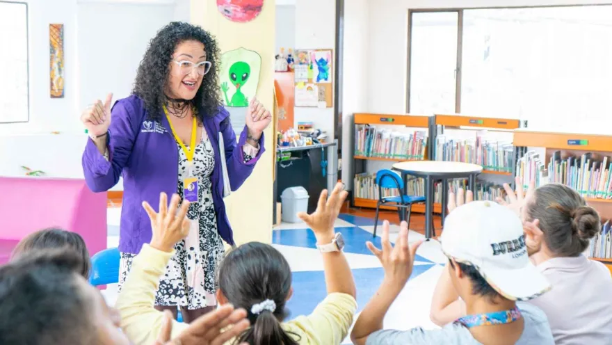 A library facilitator interacts with a group of children raising their hands in a colorful space that encourages reading and learning.