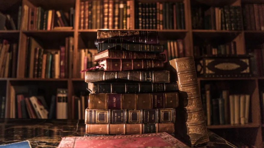 Stack of antique books on a table, with shelves full of volumes in the background of a warm library.