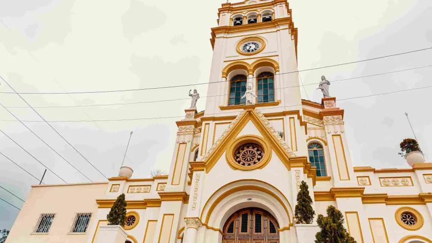 Close-up of the facade of a light brown church with touches of yellow. There are some small pine trees on either side.