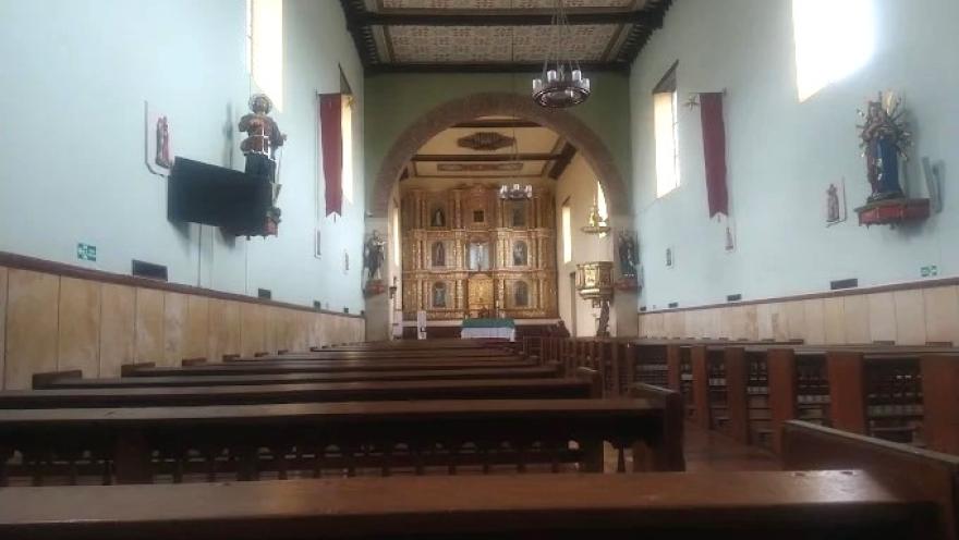Interior of a church with wooden pews, white walls, and a gilded altar at the back, illuminated by natural light entering through side windows.