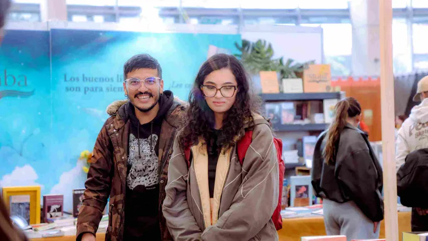 Two visitors pose and explore a book display booth at a literary fair, in the middle of a lively cultural atmosphere packed with readers, publishers, and reading-centered activities.
