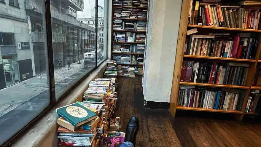 View from a bookstore with shelves and stacks of books next to a window, while a person rests looking out at the street.