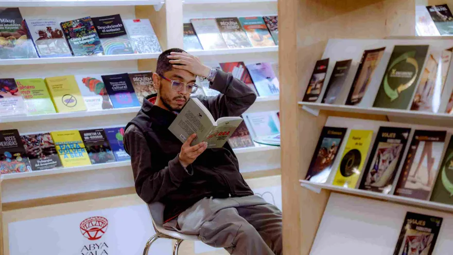 A visitor rests and reads a book intently in front of shelves filled with new releases and editorial publications at a literary fair.