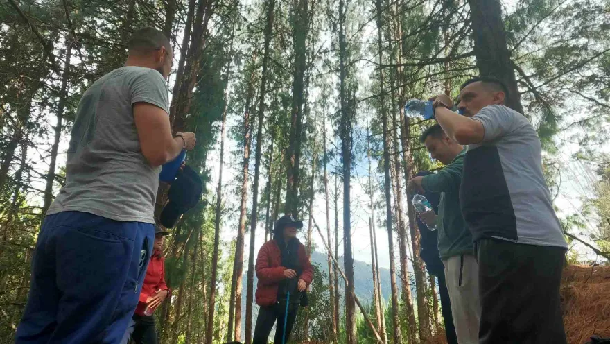 Grupo de personas en un bosque hace una pausa durante una caminata, hidratándose y conversando entre árboles altos y luz natural filtrada.