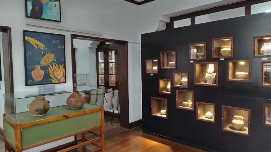  Sala de exposición de un museo arqueológico con piso de madera, vitrinas de vidrio y nichos iluminados en la pared que exhiben piezas de cerámica precolombina. En las paredes se observan pinturas de temática indígena y, al fondo, una puerta comunica con otra sala del museo.