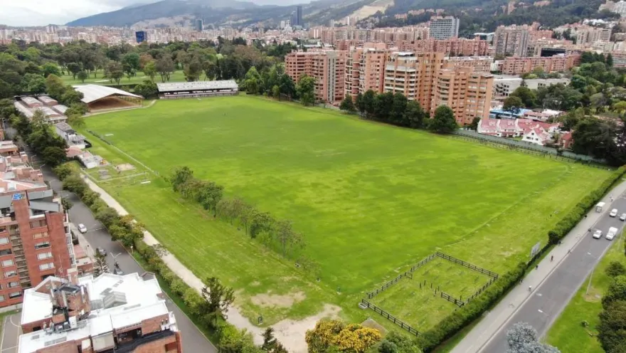 Vista aérea de una amplia cancha de césped rodeada de árboles, edificios residenciales y vías urbanas, con los cerros orientales al fondo.