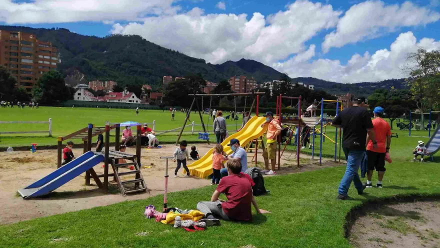 Niños y familias disfrutan de una zona de juegos infantiles y amplias áreas verdes en un parque, con los cerros orientales y un cielo parcialmente nublado al fondo.