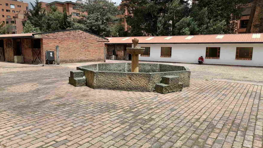Patio adoquinado con una fuente de piedra en el centro, rodeado por edificaciones de estilo tradicional, árboles altos y edificios residenciales al fondo.