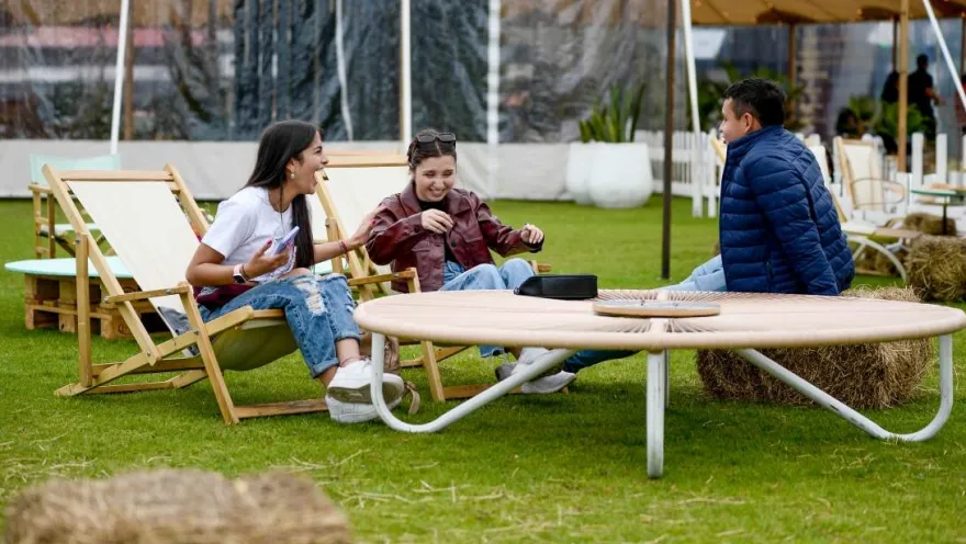 Tres personas conversan y comparten un momento de esparcimiento en sillas reclinables sobre una zona verde, junto a mesas y espacios al aire libre para eventos.