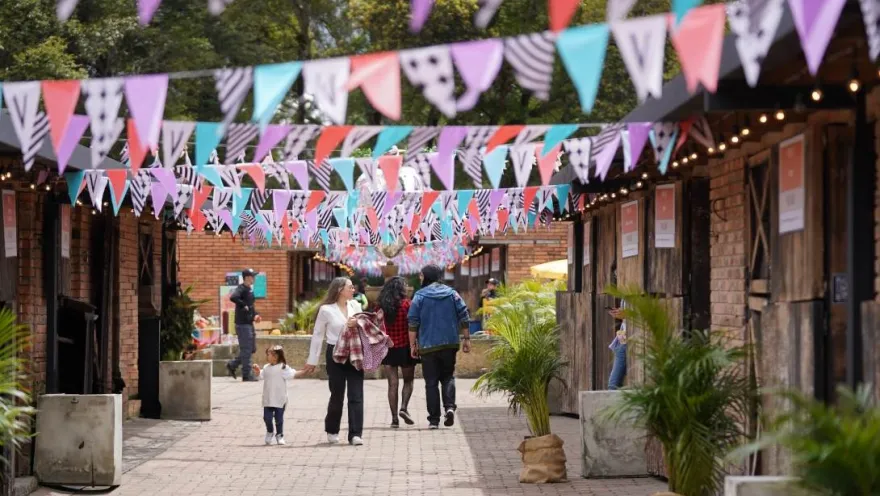 Pasillo peatonal decorado con banderines de colores, donde familias y visitantes recorren una feria al aire libre entre locales y zonas verdes.