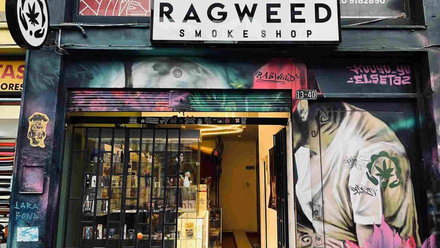 The facade of a shop with the sign "Ragweed Smokeshop," surrounded by urban art murals and graffiti. The entrance, with a black gate and illuminated display cases, showcases various products inside.