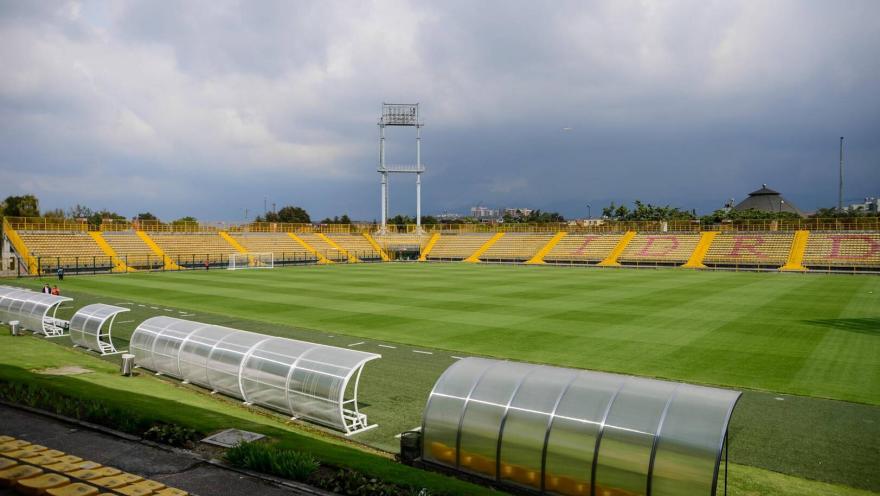  Vista panorámica del Estadio de Techo con la cancha de césped, graderías amarillas y los bancos de suplentes en primer plano bajo un cielo nublado.