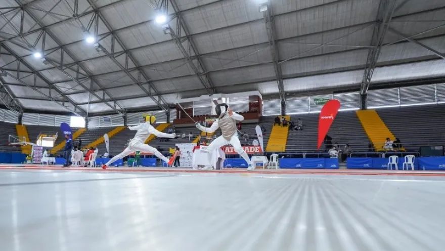 Athletes compete in a fencing match inside the Palacio de los Deportes in Bogotá, on a piste surrounded by stands and competition areas.