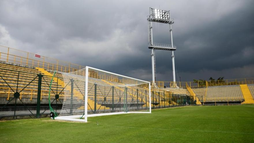 Arco de fútbol sobre la cancha del Estadio de Techo, con graderías amarillas, torre de iluminación y un cielo oscuro y nublado al fondo.