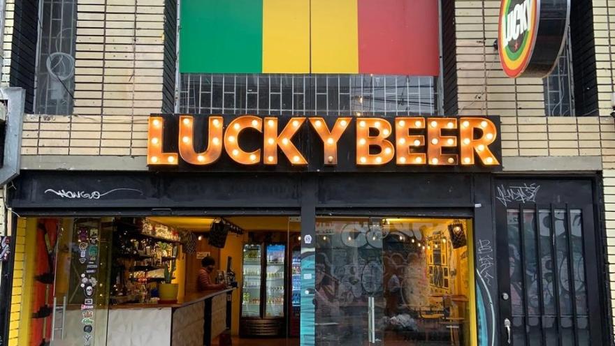 The facade of Lucky Beer with an illuminated sign, a green, yellow, and red flag above the entrance, and a bar visible through its glass doors.