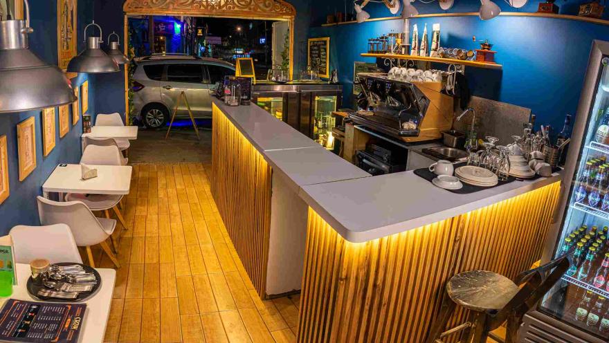 The interior of a coffee shop with an illuminated wooden counter, a coffee machine, white tables, and blue-toned decor, seen from the entrance at night.
