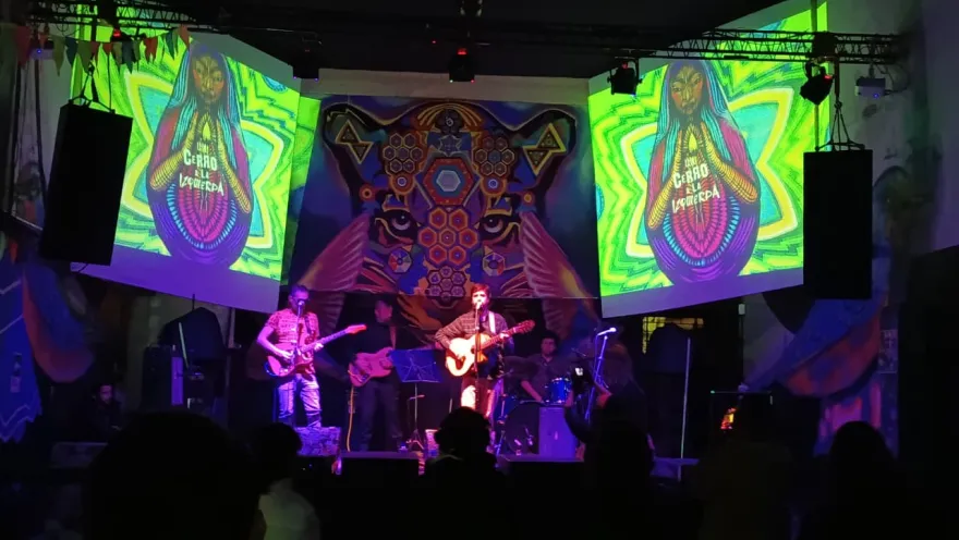 Escenario con una banda tocando en vivo frente al público, acompañado de proyecciones coloridas y un mural artístico de fondo en un recinto de conciertos.