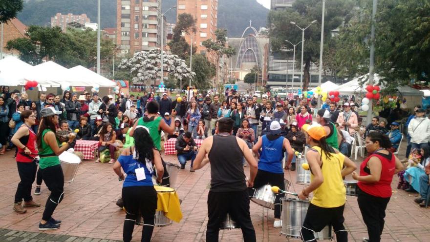 A group of artists performs a musical presentation with drums in front of numerous spectators in an urban park in Bogotá.