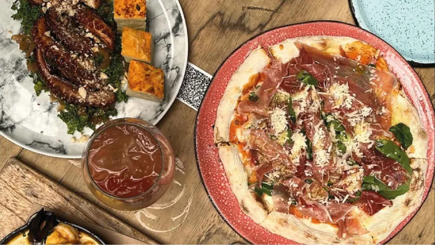 Vista superior de una mesa de madera con una pizza de jamón, un plato de pulpo, un cóctel y aperitivos variados.