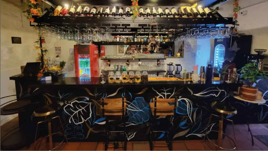 Un bar con una encimera de madera oscura pintada con hojas azules y blancas, y estantes llenos de botellas de alcohol.