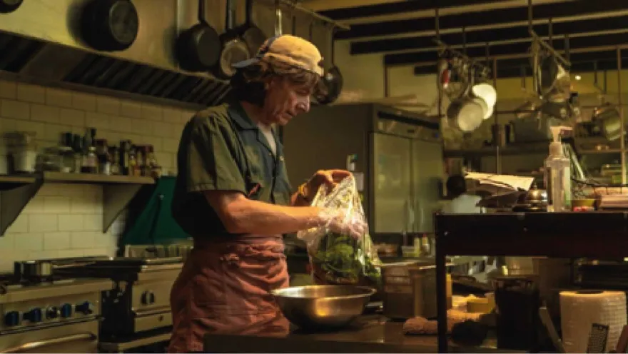 Un chef con una gorra de béisbol y un delantal de cocina prepara verduras en una bolsa de plástico en una cocina rústica.