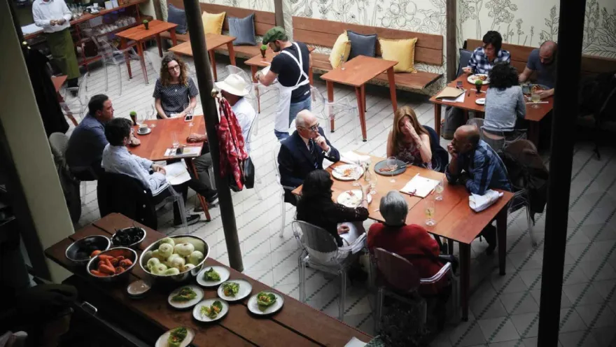 Vista aérea de un patio de restaurante lleno de gente cenando en mesas de madera, con una cocina abierta visible.