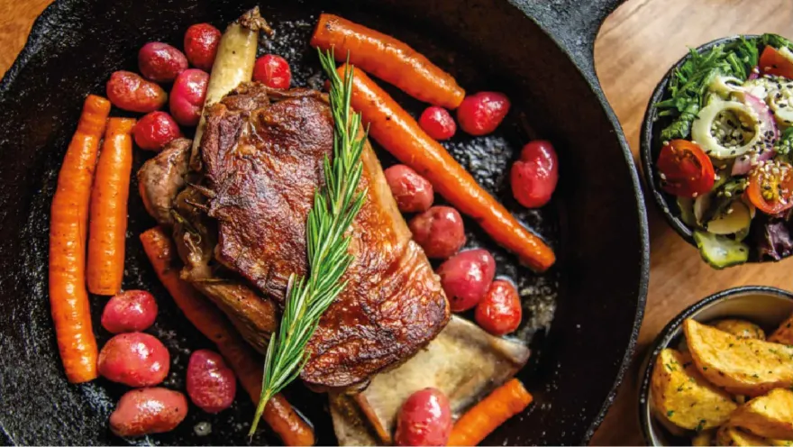  Cacerola de hierro con un corte de carne, romero, zanahorias y papas, acompañada de ensalada y papas fritas.