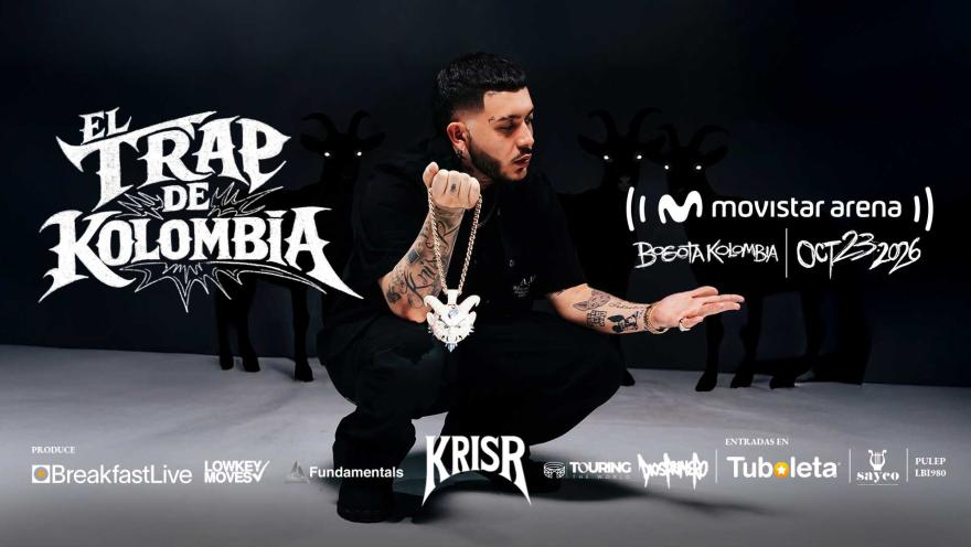 El artista Kris R agachado con collar de cabeza de cabra, rodeado de sombras de cabras al fondo.