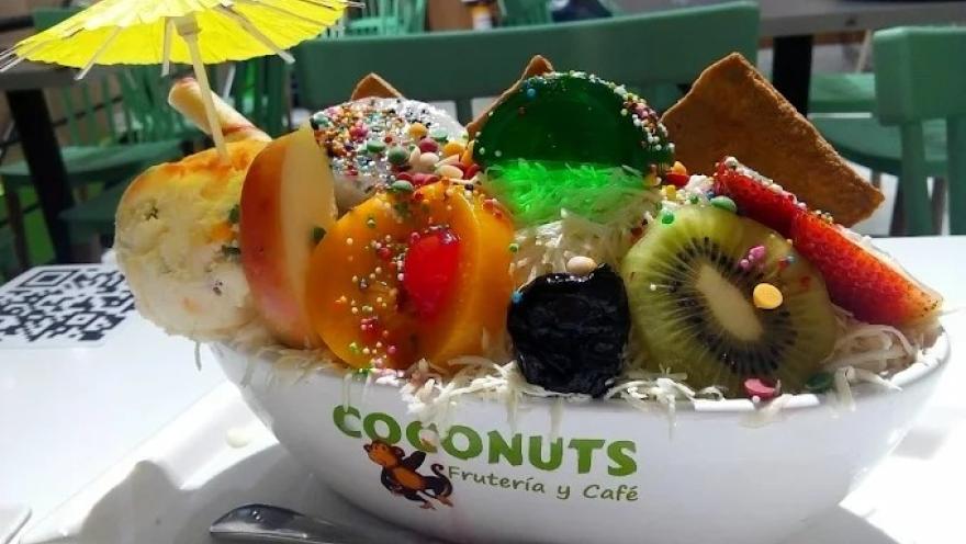 Copa de frutas tropicales con helado, gelatina, coco rallado, grageas y salsa dulce, servida en un recipiente de la frutería.