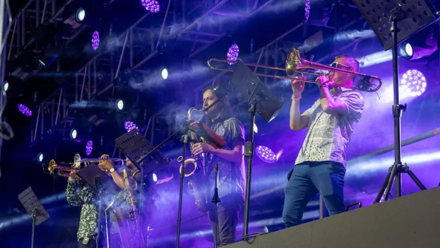 Músicos en escenario tocan trombón y saxofón bajo luces moradas durante un concierto en vivo.
