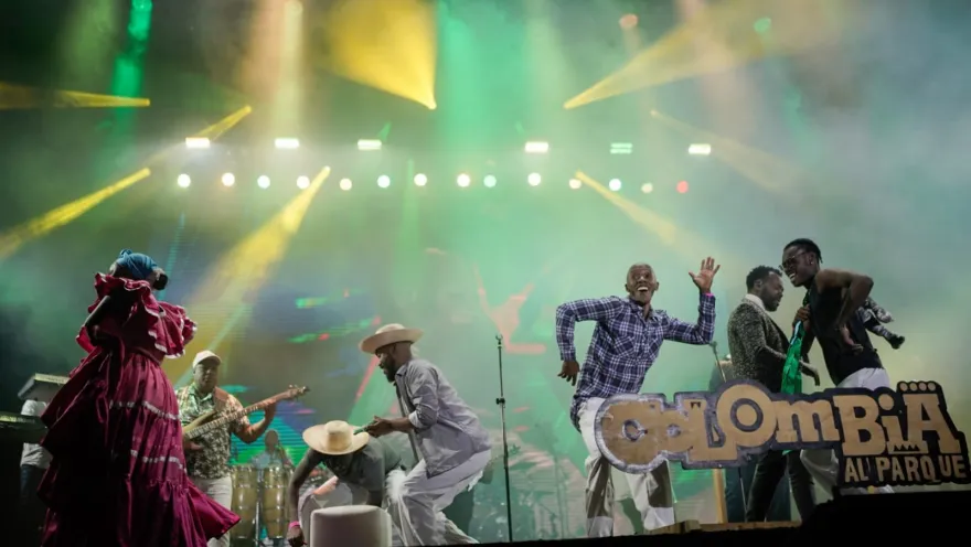 Grupo de músicos y bailarines se presenta en un escenario con luces de colores durante un concierto.