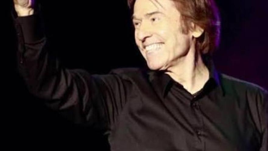 Cantante en el escenario, vestido de negro, sonriendo y saludando al público mientras sostiene un micrófono en una mano.