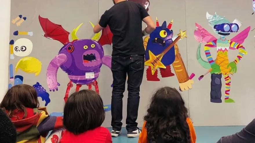 A group of children watches an artist create a colorful piece featuring fantastical characters and monsters of different shapes, sizes, and colors in a space dedicated to children's art and creativity.
