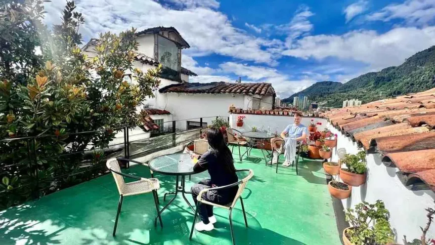 Terraza con mesas y sillas al aire libre, rodeada de plantas y con vistas a los tejados tradicionales y las montañas de Bogotá.