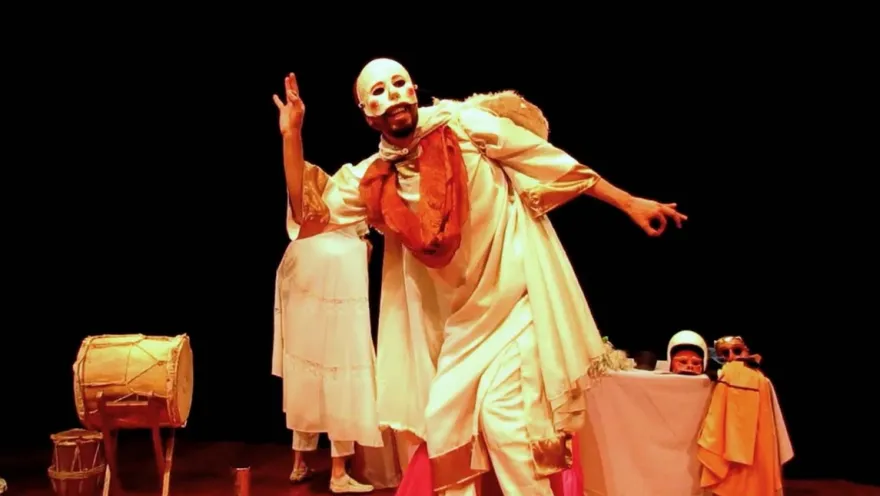 An artist wearing a white mask and traditional costume stars in a theatrical scene full of expression, music, and symbolism, accompanied by drums and other stage elements.