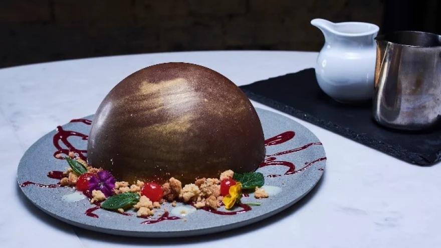 Postre de chocolate en forma de esfera, decorado con frutos, flores comestibles y salsa de frutos rojos.