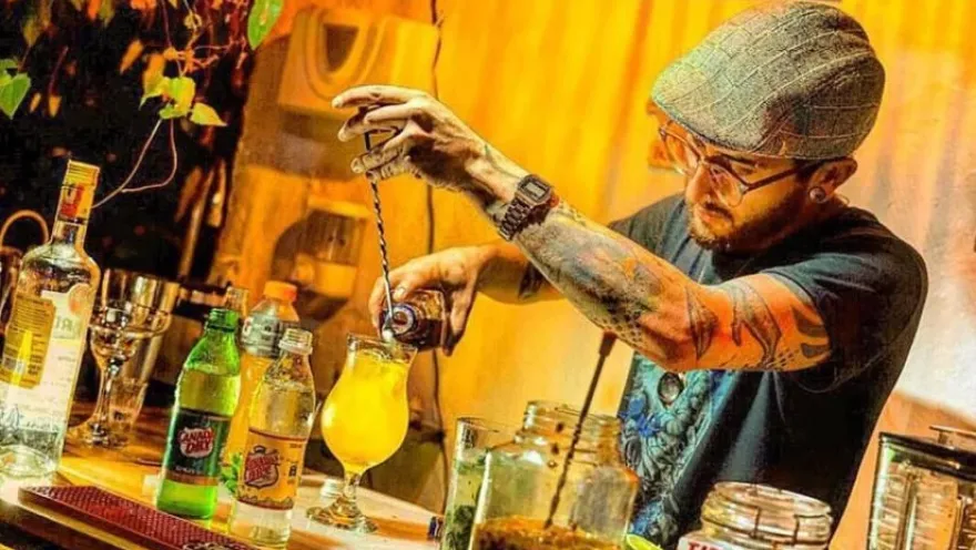 Bartender preparando un cóctel en una barra, rodeado de botellas y utensilios de coctelería en un ambiente cálido.