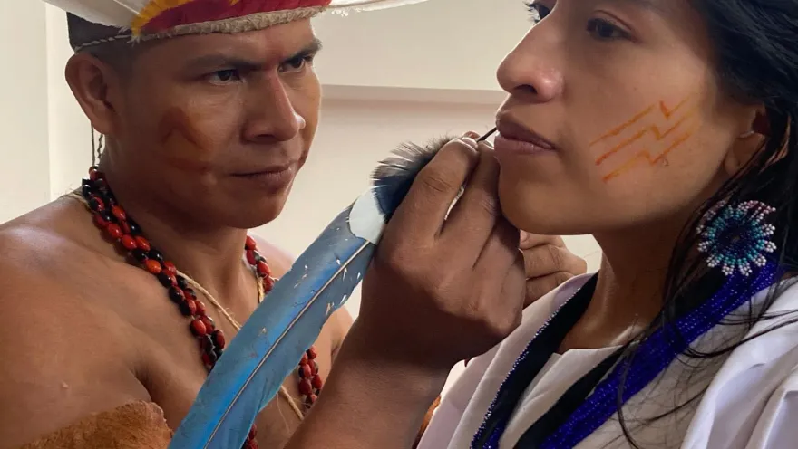 Un representante indígena realiza una pintura facial tradicional a una visitante, como parte de una experiencia de acercamiento a las costumbres y expresiones culturales de los pueblos originarios.