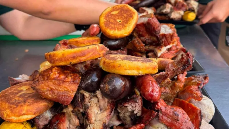 Bandeja de picada gigante repleta de carnes asadas, embutidos, papas saladas y arepas servidas al centro de la mesa.