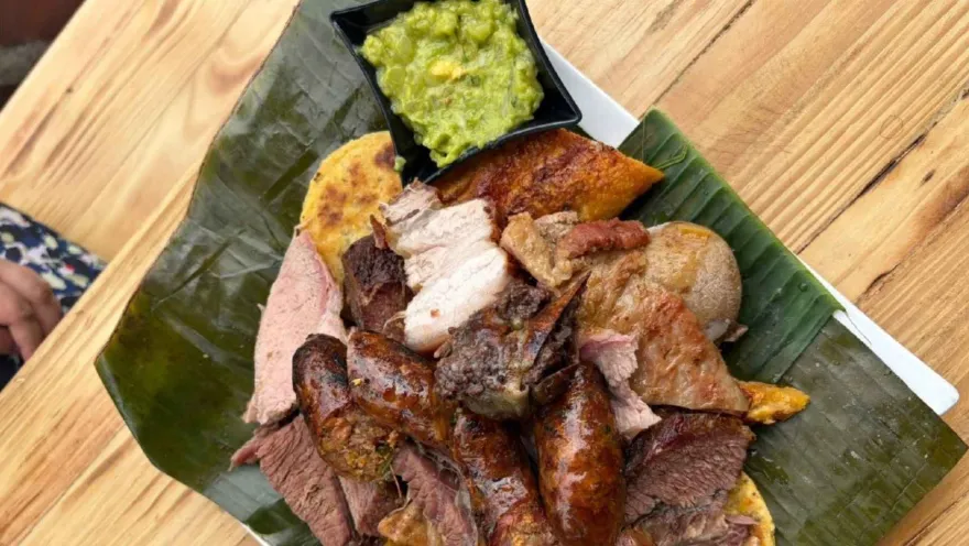Plato servido con cortes de carne asada, chorizos dorados, papa y un recipiente con guacamole sobre mesa de madera.   