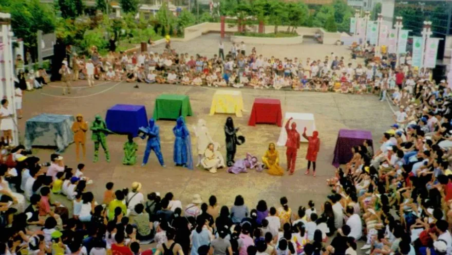 Audience gathered in a public square to enjoy an outdoor street theater performance, featuring colorfully dressed artists and an open-air staging.