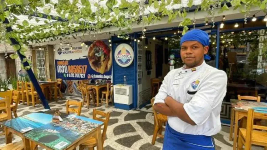 Cocinero con delantal blanco y pañuelo azul en la cabeza sonríe de pie en la terraza iluminada de un restaurante.   