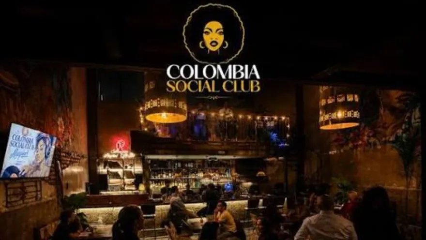 Vista panorámica del bar con ambiente tenue, clientes en las mesas y un logo dorado brillante destacado al centro.   