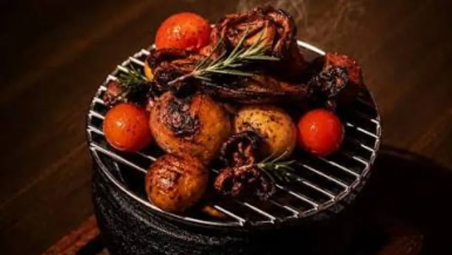 Mini anafre de hierro caliente con brochetas de carne, papas doradas, tomates rojos y romero sobre mesa de madera.   